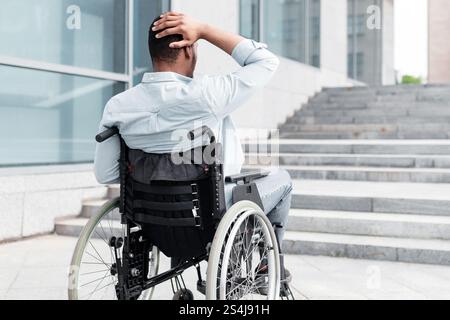 Uomo nero irritato in sedia a rotelle che non ha la possibilità di entrare nell'edificio senza rampa, all'aperto Foto Stock