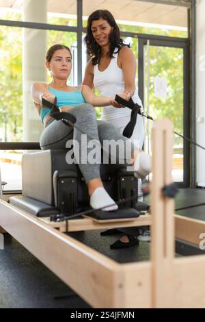 Istruttrice di pilates femminile ispanica che aiuta la giovane donna a fare esercizi sull'apparato riformatore Foto Stock