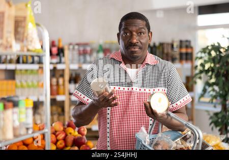 L'uomo africano sta tenendo in mano due lattine di latta di conserve, scegliendo il patè per i panini. Foto Stock