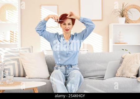 Donna matura che si taglia i capelli rossi sul divano a casa Foto Stock