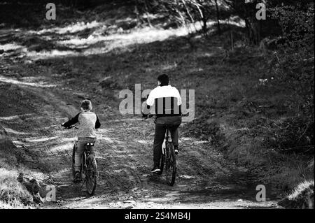 Un'immagine in bianco e nero cattura una scena drammatica di padre e figlio che pedala lungo un rustico sentiero sterrato. L'ambiente è sereno e all'aperto, emphas Foto Stock
