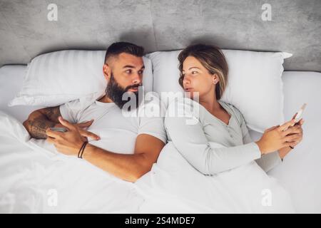 La giovane coppia felice è sdraiata a letto la mattina. Usano gli smartphone. Foto Stock