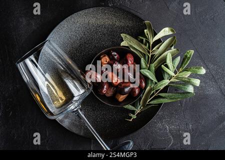 Vista dall'alto di una sistemazione sofisticata con olive greche al barbecue immerse in una ciotola nera con foglie di alloro fresche, accompagnate da un bicchiere di bianco secco Foto Stock