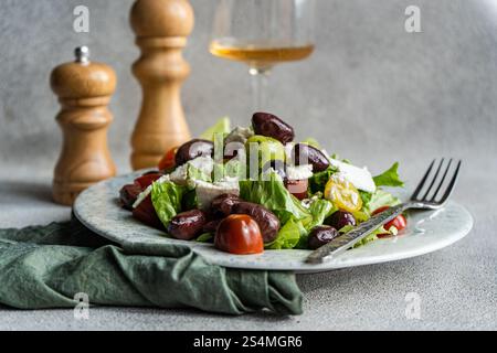 Una vivace insalata greca con pomodori, lattuga, olive rosse, formaggio feta e olio d'oliva Un bicchiere di vino bianco freddo completa il pasto salutare Foto Stock