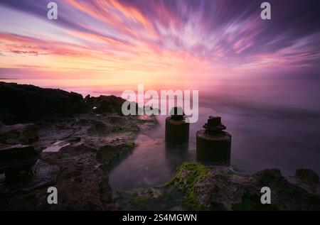 Un tramonto ipnotico con vivaci sfumature che si riflettono sulle acque calme, sulle rocce costiere e sulle antiche rovine o strutture, creando una tranquilla e suggestiva S Foto Stock