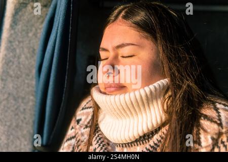 Un ritratto ravvicinato di una giovane e premurosa donna latina colombiana, crogiolata alla luce calda del sole con gli occhi chiusi, che indossa una grossa swea lavorata a maglia Foto Stock