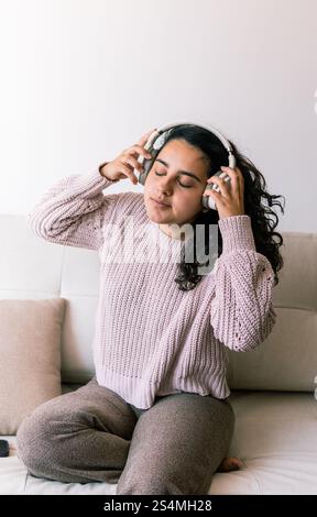Una giovane donna latina con i capelli ricci si rilassa sul divano di casa, indossando un comodo maglione e cuffie, ama ascoltare musica con gli occhi clos Foto Stock