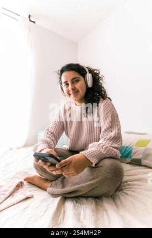 Una giovane donna latina che si diverte a casa, indossa le cuffie e utilizza uno smartphone mentre si siede su un comodo letto, interni luminosi e informali Foto Stock