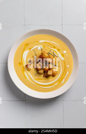 Un primo piano di cremosa zuppa di curry di cavolfiore guarnita con crostini croccanti e rosmarino presentati su un piatto di ceramica su uno sfondo piastrellato bianco, evo Foto Stock