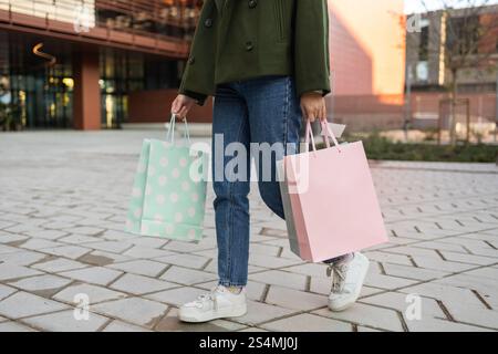 Una donna con un cappotto verde e un jeans porta borse della spesa pastello cammina su un marciapiede a motivi geometrici, mostrando una piacevole esperienza di shopping urbano con spirito Foto Stock