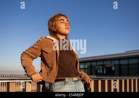 Una giovane donna di corsa mista con giacca marrone e jeans si erge accanto a una ringhiera, godendosi il sole, il cielo blu brillante e l'architettura moderna suggeriscono un Foto Stock