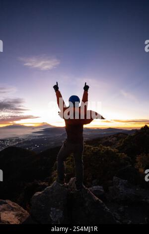 Vista posteriore di un uomo irriconoscibile che festeggia sulla cima di una montagna durante l'alba, con le mani alzate e il paesaggio della città visibile in lontananza. Foto Stock