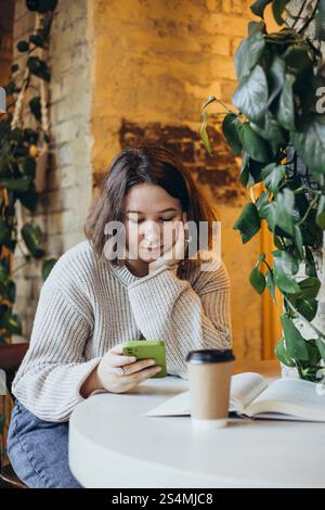 Una giovane donna in un maglione accogliente siede al tavolo di un bar libreria, scorrendo sul suo smartphone un libro aperto e una tazza di caffè sono di fronte a lei, surrogato Foto Stock