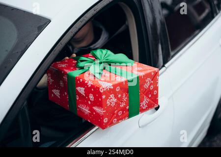 Una persona consegna un regalo natalizio avvolto in rosso con un fiocco verde dal finestrino di un'auto. La scena cattura un momento di donazione, mettendo in risalto la gioia e le vacanze Foto Stock