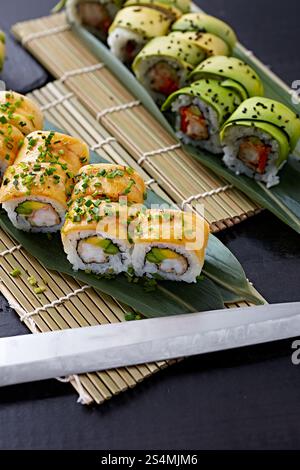 Varietà di sushi panini elegantemente posizionati su un tappeto di bambù, con condimenti di avocado e pesce, guarniti con semi di sesamo e cipolle verdi. Foto Stock