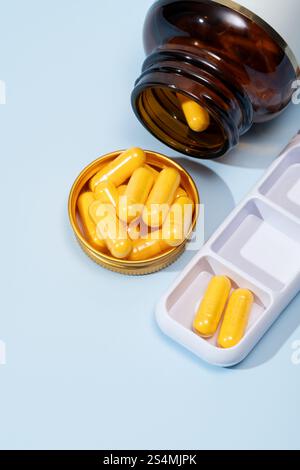 Vista dall'alto di una bottiglia color ambra scuro che versa capsule gialle in un coperchio dorato, accanto a un organiser per pillole bianco su sfondo blu. Foto Stock
