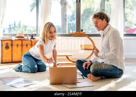 Un uomo e una donna seduti sul pavimento di casa, impegnati in attività di lavoro e pianificazione. L'uomo e la donna stanno guardando un portatile e dei documenti mentre si' Foto Stock