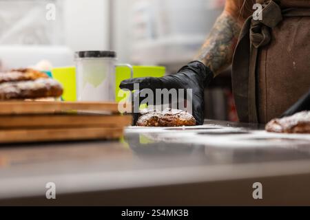 Un fornaio esperto con le braccia tatuate e i guanti dispone di dolci appena sfornati su un bancone. L'atmosfera calda e invitante riflette un vero e proprio IT Foto Stock