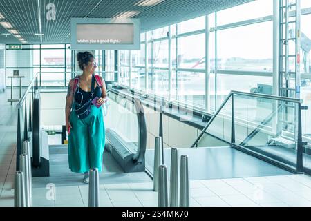 Un viaggiatore si trova su una scala mobile in un aeroporto luminoso, con in mano Un passaporto, Un cartello sopra vi augura un volo sicuro le pareti di vetro rivelano gli aerei all'esterno Foto Stock