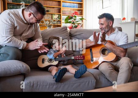 Una coppia gay ama trascorrere momenti di qualità con il suo giovane figlio, suonando le chitarre nell'accogliente soggiorno. Condividono sorrisi e applausi, promuovendo il creat Foto Stock