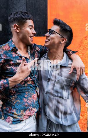 Una allegra coppia gay latina condivide un momento giocoso, sorridendo l'un l'altro contro una vivace parete arancione e nera, mostrando affetto e connessione Foto Stock