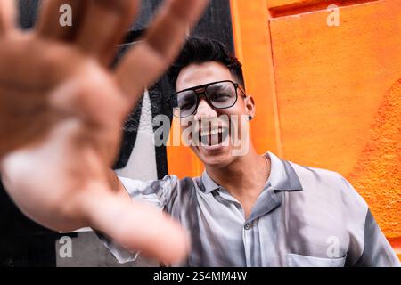 Un giovane uomo che indossa occhiali e una camicia elegante sorride con gioia, estendendo la mano verso la fotocamera. Il vivace sfondo arancione migliora il vivo Foto Stock