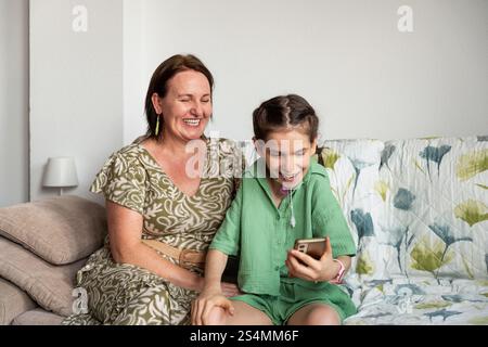Una scena gioiosa di una madre e di sua figlia disabile, che ha una tracheostomia, seduto dentro e condividendo momenti di felicità guardando un intelligente Foto Stock