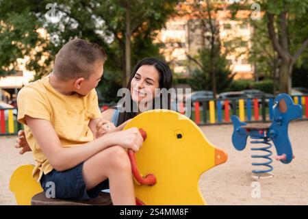 Una madre amorevole e suo figlio con paralisi cerebrale condividono un momento di gioia al parco giochi, evidenziando l'amore, il sostegno e l'inclusione in un vivace outdoo Foto Stock