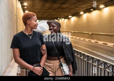 Una coppia multietnica LGBT condivide un momento di gioia mentre passeggia attraverso un tunnel urbano illuminato. Camminano braccio in braccio, irradiando felicità e c Foto Stock