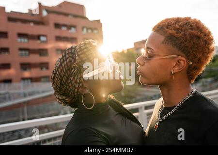 Una coppia multietnica LGBT abbraccia in un momento tenero al tramonto. Indossano eleganti occhiali da sole e abbigliamento casual. Il sole caldo crea un bel bagliore Foto Stock