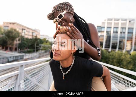Una gioiosa coppia multietnica LGBT condivide momenti divertenti all'esterno. Si godono la presenza reciproca su un moderno ponte cittadino, circondato da paesaggi urbani Foto Stock