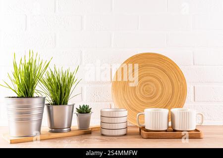 Un ambiente contemporaneo da cucina caratterizzato da un piatto rotondo in legno, tazze a righe bianche su un vassoio e varie piante verdi in vaso contro un wal in mattoni bianchi Foto Stock