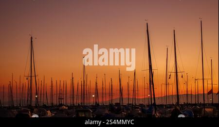 Serena vista del tramonto sul lago Leman con diversi alberi di barca a vela che si stagliano contro un vivido cielo arancione, catturato a Losanna. Foto Stock