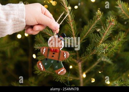 Uno scatto ravvicinato di una mano di un bambino corta e irriconoscibile appesa a un ornamento di un uomo di pan di zenzero su un albero di Natale splendidamente illuminato, catturando l'essenza Foto Stock