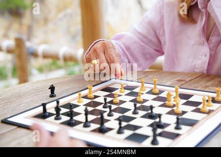 Primo piano di mani che muovono pezzi di scacchi durante una partita giocata all'esterno su un tavolo di legno, il tabellone mostra una posizione strategica a metà partita in una giornata di sole Foto Stock