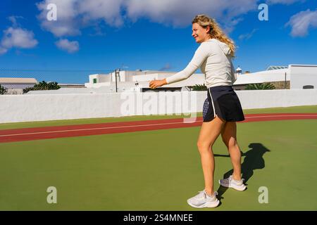 Una giovane donna dai capelli biondi ricci gioca attivamente a tennis, catturata in un profilo laterale mentre si prepara a colpire un colpo al rovescio. Sta indossando una W Foto Stock