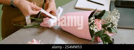 Banner del sito Web con un inriconoscibile fiorista che strizza fiorista con un nastro di sating intorno a una composizione floreale in carta da imballaggio rosa pastello Foto Stock