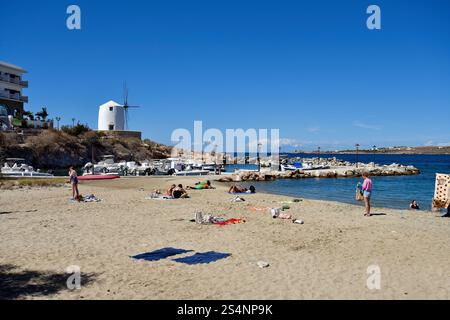 Paros, Grecia - 17 settembre 2024: Persone sconosciute sulla spiaggia di Parakia con un piccolo porto di pescatori e un mulino a vento che viene utilizzato come caffè Foto Stock