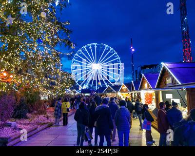 I visitatori passeggiano attraverso il vivace mercato di Natale in Place de Jaude, Clermont-Ferrand, con bancarelle illuminate, una ruota panoramica gigante e un vib Foto Stock
