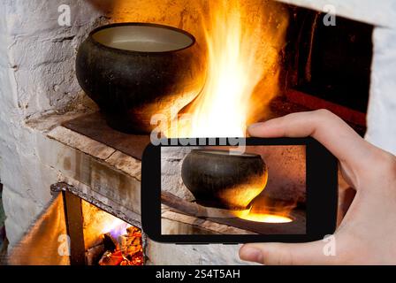fotografare il concetto di cibo - il turista scatta una foto del focolare del fuoco della stufa russa e del vecchio vaso di ghisa con forchetta da forno su smartphone, Foto Stock