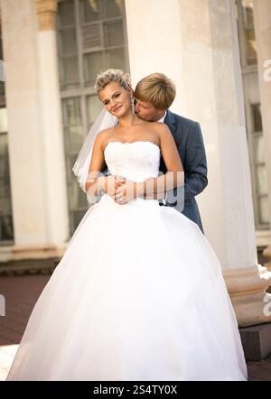 Bellissimo giovane sposo che abbraccia la sposa sorridente da dietro per strada Foto Stock