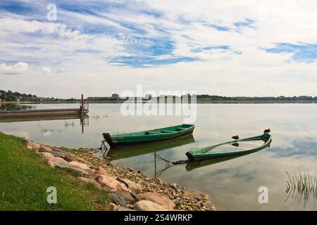 Calmo lago con due piccole barche verdi su una riva rocciosa con anatre sotto un cielo tranquillo, che riflettono la serenità della natura. Foto Stock