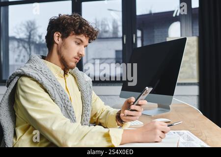 Il giovane con i capelli ricci usa il suo smartphone a una scrivania in uno spazio di lavoro moderno. Foto Stock