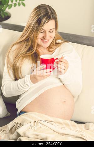 Ritratto di una donna incinta sorridente e felice seduta sul divano con una tazza di tè Foto Stock