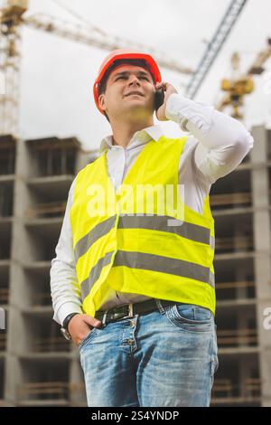 Giovane ingegnere sorridente in elmetto e giubbotto di sicurezza che parla al telefono sul cantiere Foto Stock