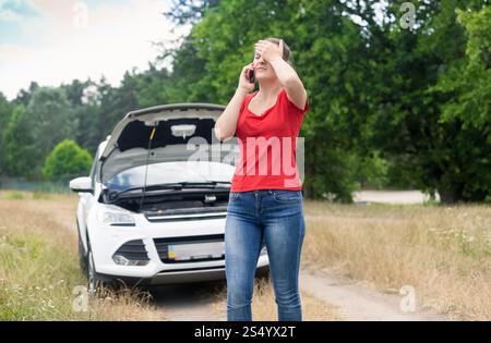 Donna triste in piedi davanti a un'auto rotta e chiamata al cellulare per chiedere aiuto Foto Stock