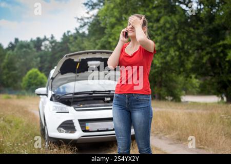 Donna in piedi davanti a un'auto rotta sulla strada rurale e che chiama al cellulare per chiedere aiuto Foto Stock