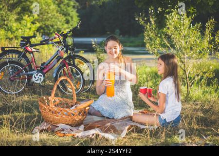 Madre che fa un picnic al fiume con la figlia. La madre versa il succo d'arancia nella tazza delle figlie Foto Stock