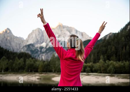Una donna con una giacca rosa brillante alza le braccia in vittoria, davanti a uno splendido paesaggio di montagna circondato da una vegetazione lussureggiante. Foto Stock
