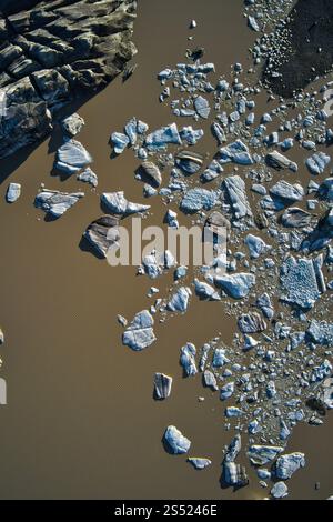 Vista aerea dei blocchi di ghiaccio galleggianti in una laguna glaciale, circondata da acque fangose e terreni accidentati, catturando la bellezza selvaggia dell'Islanda Foto Stock
